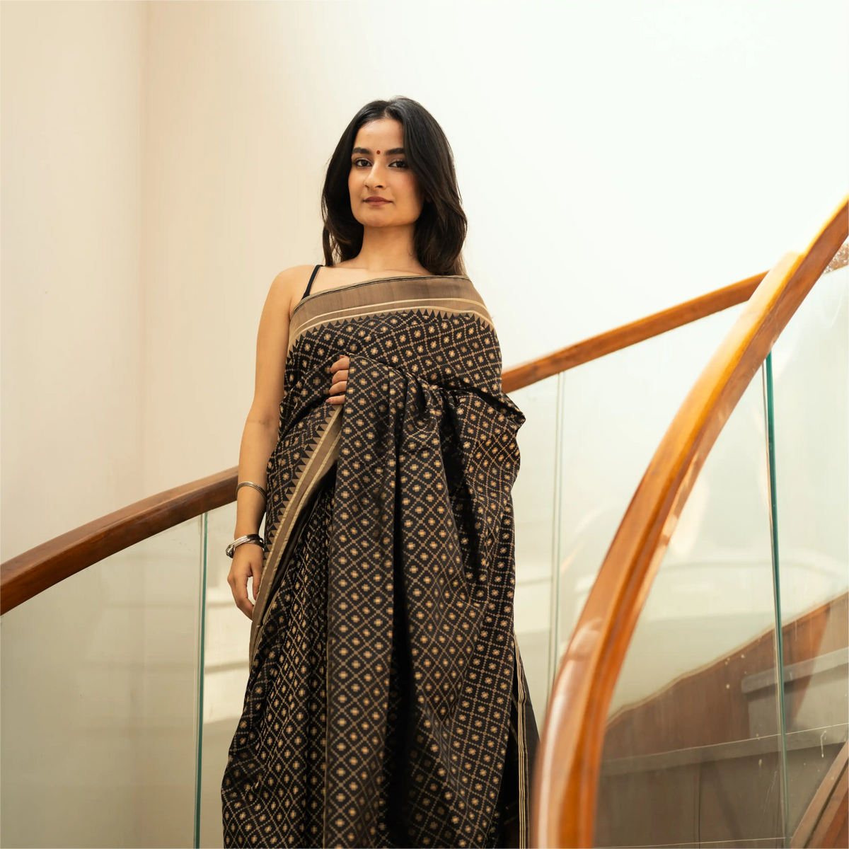 A model posing in an elegant black saree with a gold-toned ethnic pattern, standing on a staircase with a wooden railing.