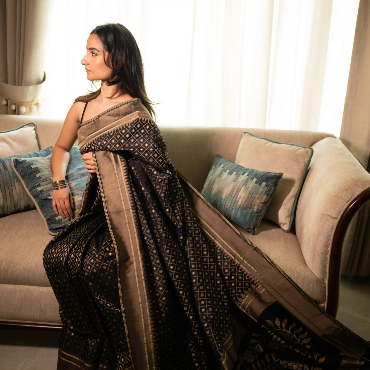 A woman wearing an elegant black Banarasi silk saree with intricate gold zari work, sitting gracefully on a beige sofa in a softly lit room.
