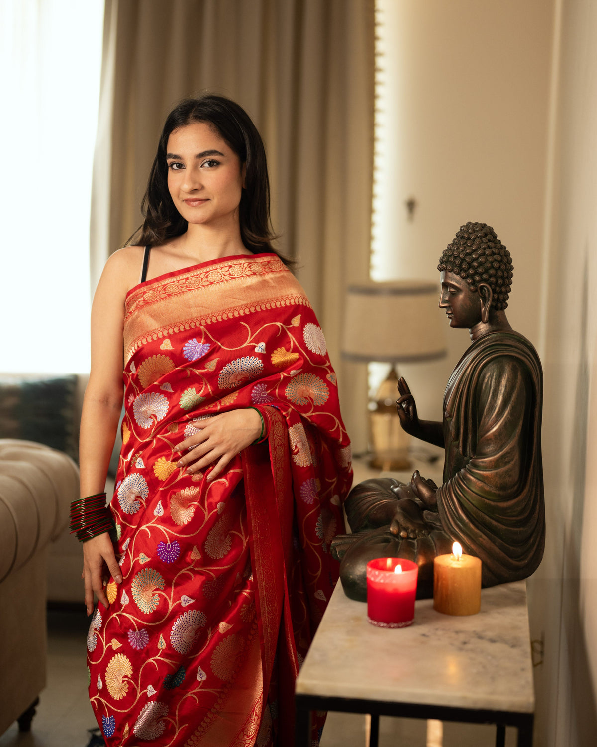 A woman wearing a vibrant, elegant red handwoven Banarasi silk saree with intricate gold zari work, standing indoors near a decorative Buddha statue