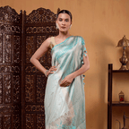 Model draped in a light blue and white celestial-toned Aurora Borealis Katan Silk Saree featuring a horizontal gold zari and resham border, posing against a wooden backdrop.