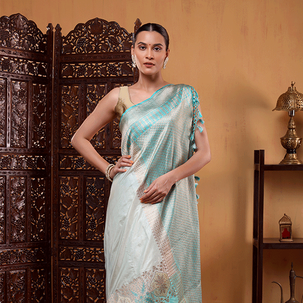 Model draped in a light blue and white celestial-toned Aurora Borealis Katan Silk Saree featuring a horizontal gold zari and resham border, posing against a wooden backdrop.