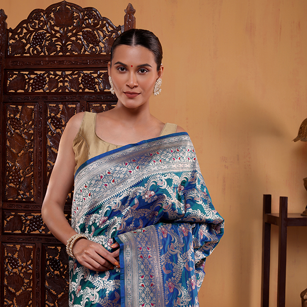 Model draped in a blue Azure Meenakari Majesty Katan Silk Saree with handwoven Shikargah patterns and a gold border, posing against a wooden backdrop.