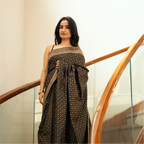 Model wearing a Black Handwoven Katan Silk Saree with Gold Zari floral work and a broad border, standing on a staircase.
