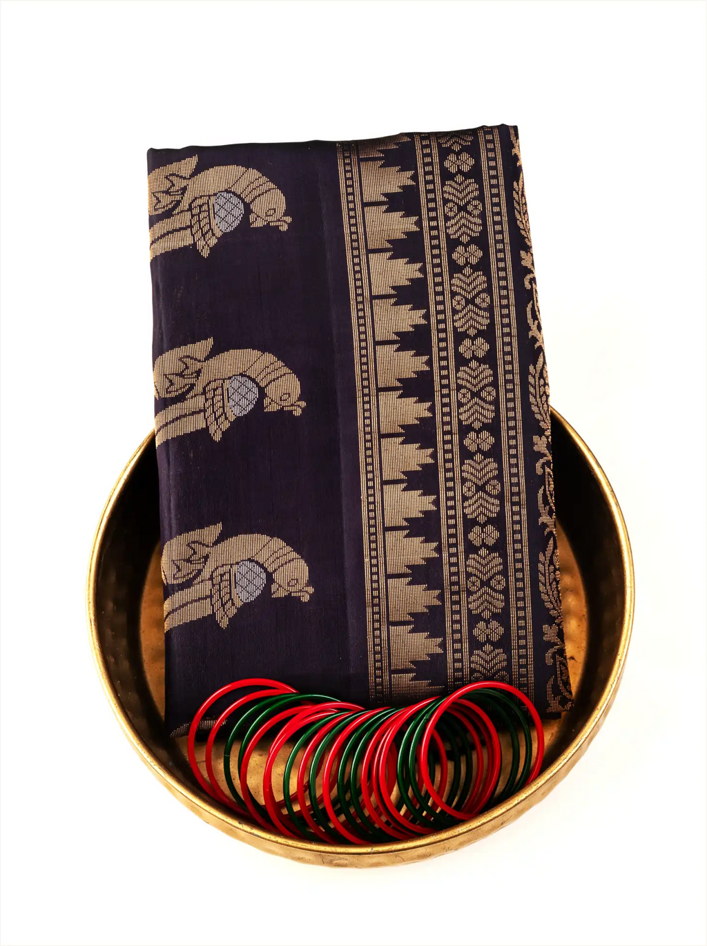 Neatly folded deep black Patola silk saree with gold woven elephant and bird motifs on the pallu, placed inside a traditional brass bowl alongside vibrant red and green glass bangles.