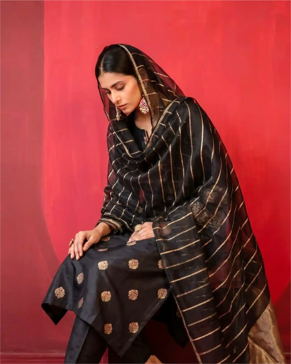 A model wearing a premium black pure Banarasi silk suit with gold zari motifs and a matching sheer Kora dupatta against a red background.