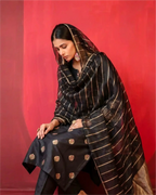 A model wearing a premium black pure Banarasi silk suit with gold zari motifs and a matching sheer Kora dupatta against a red background.