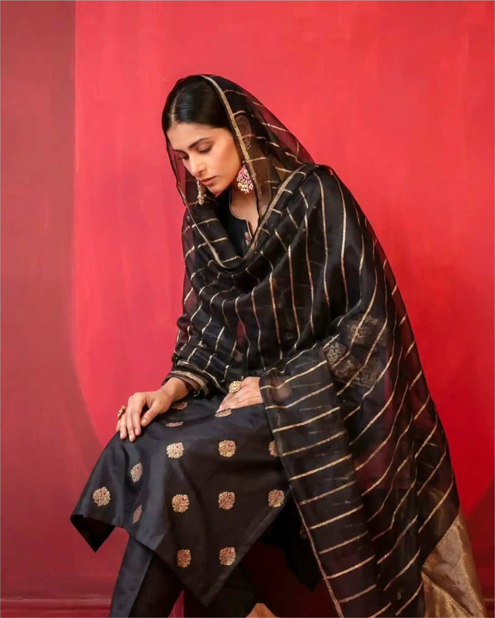 A model wearing a premium black pure Banarasi silk suit with gold zari motifs and a matching sheer Kora dupatta against a red background.