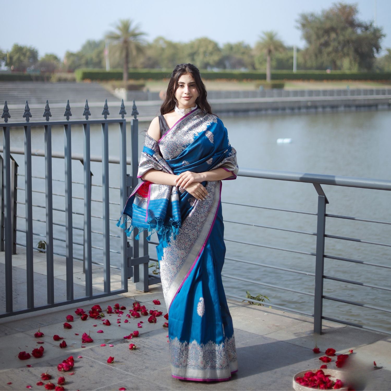 A woman standing by a body of water, wearing a blue and silver Pure Tussar Silk saree with a decorative silver border, surrounded by rose petals on the ground.