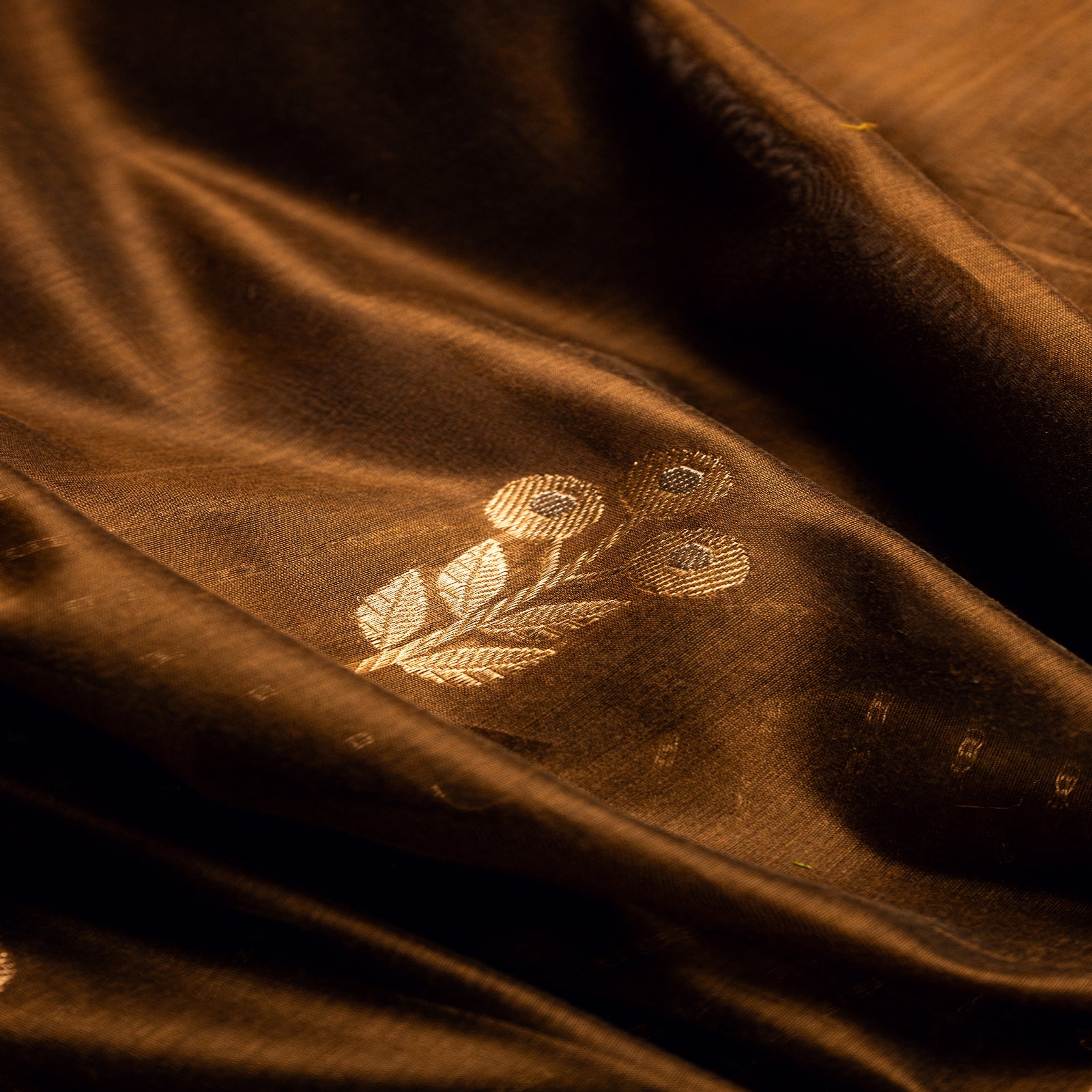 Close-up of the pleated earthy brown Alfi silk fabric, showing the fine weave and detailed gold zari floral buta motifs.