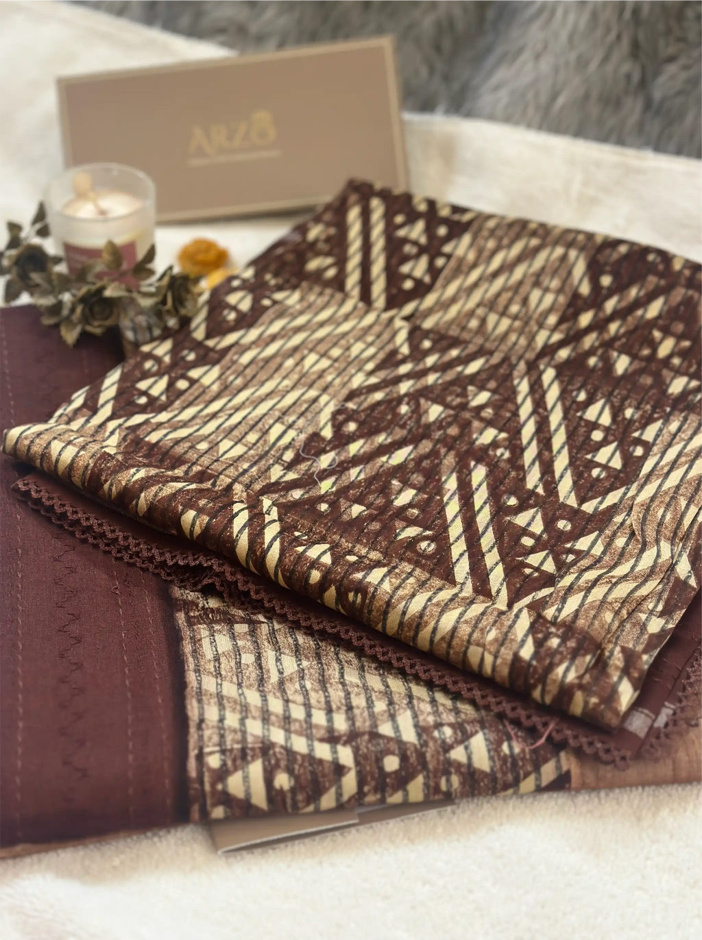 Folded brown cotton fabric with a geometric print, displayed with an Arzo-branded gift box and a small candle.