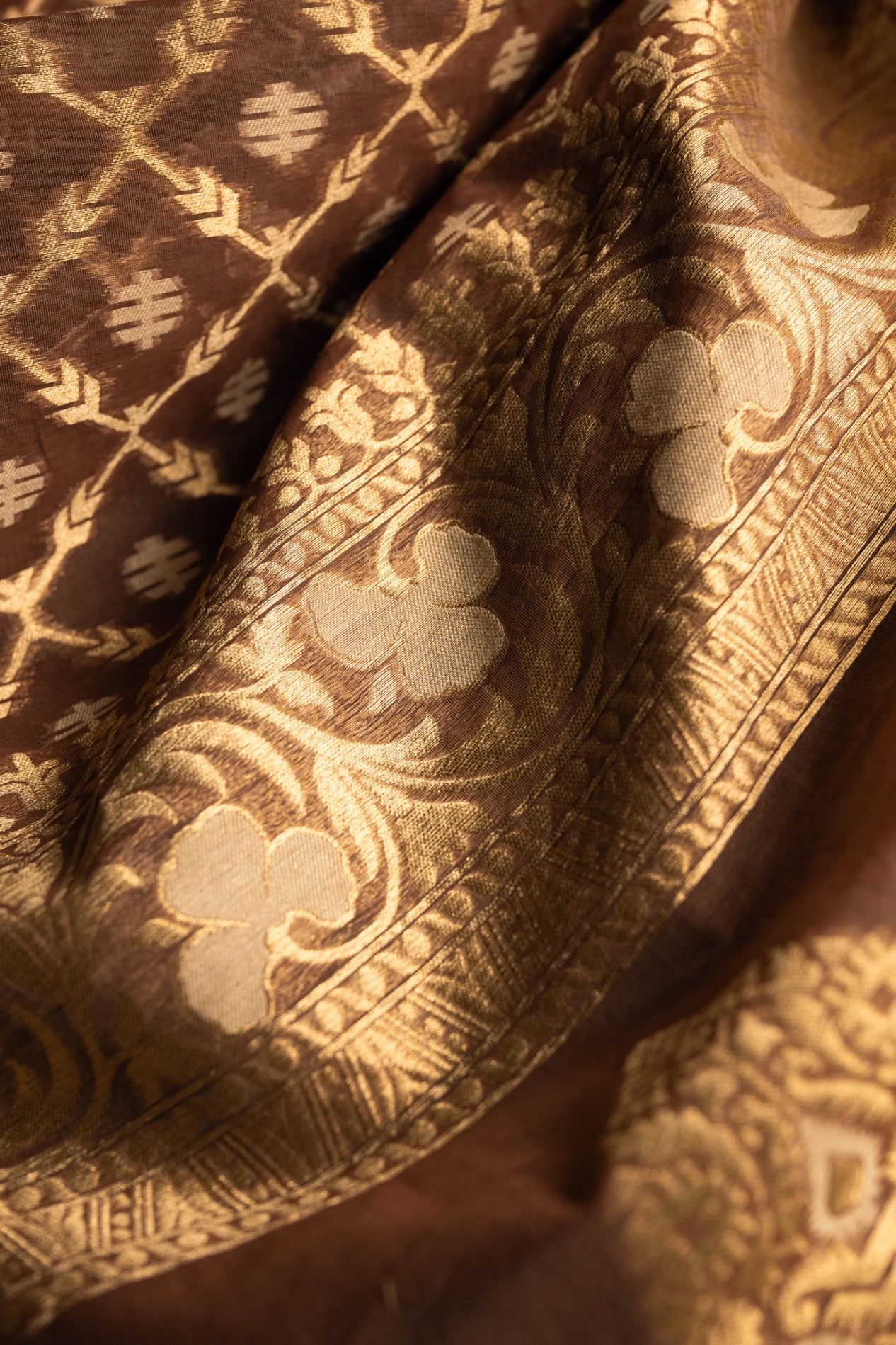 Close-up of the pleated folds of an earthy brown Jamdani silk cotton saree, showcasing the intricate gold zari work and the soft, premium texture of the hand-loomed fabric.