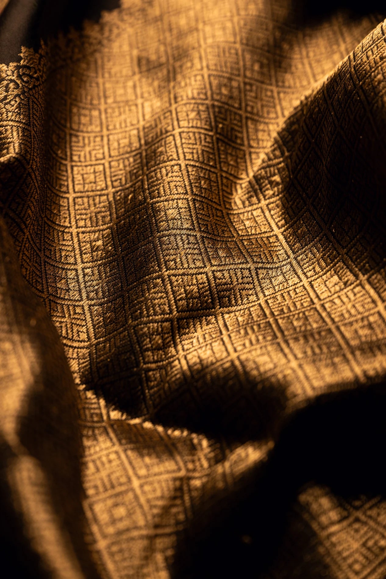 Close-up of the textured folds of an earthy brown Jamdani silk cotton saree showcasing handwoven gold zari geometric grid patterns and a rich border.