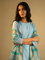 A woman posing in a light blue traditional unstitched Chanderi cotton silk suit, featuring gold bootas and a draped Rangkaat Kora dupatta with gold zari borders.