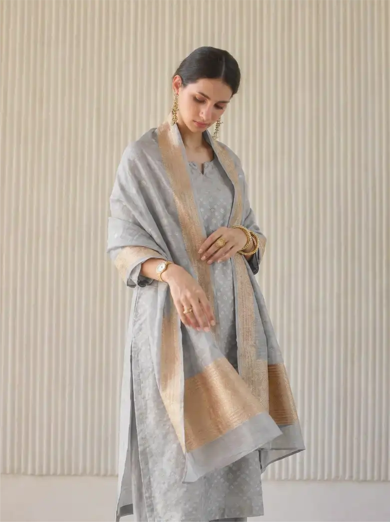 A woman posing in a premium gray unstitched Cotton Alfi suit featuring a woven texture, elegantly draped with a matching beige and gray Banarasi style borders dupatta.