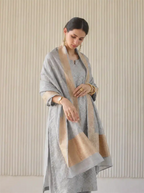 A woman posing in a premium gray unstitched Cotton Alfi suit featuring a woven texture, elegantly draped with a matching beige and gray Banarasi style borders dupatta.