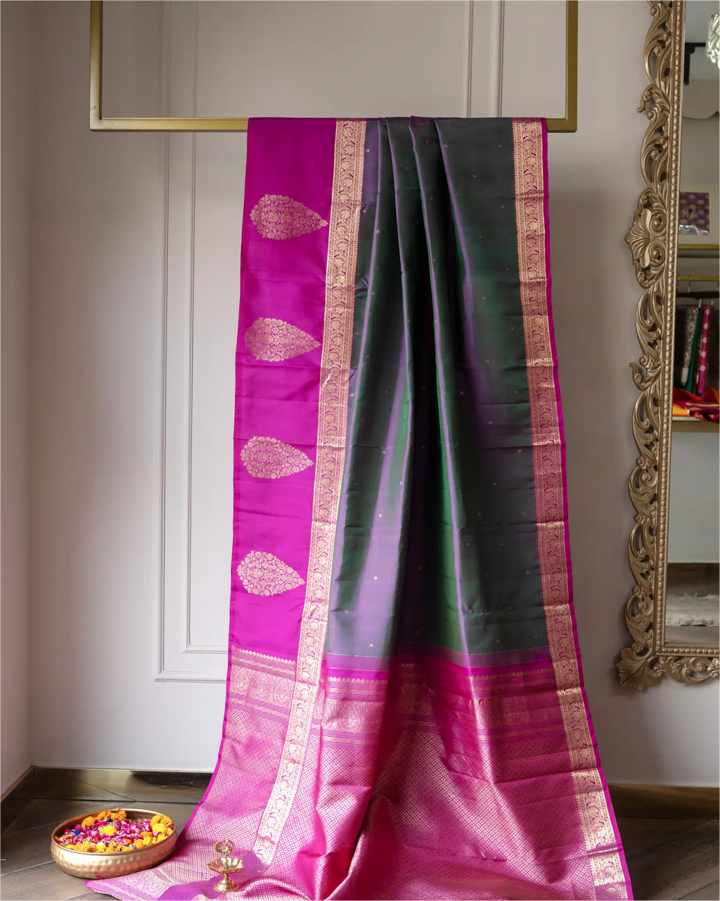 A draped silk saree with a magenta-pink body, dark green border, and ornate gold zari work, displayed indoors against a white paneled wall.