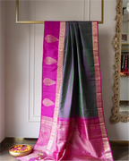 A draped silk saree with a magenta-pink body, dark green border, and ornate gold zari work, displayed indoors against a white paneled wall.