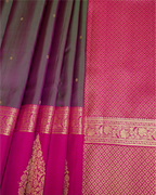 A close-up of a richly patterned silk saree, showing a dark plum purple body with small gold motifs, a bright magenta-pink border, and detailed gold zari brocade work