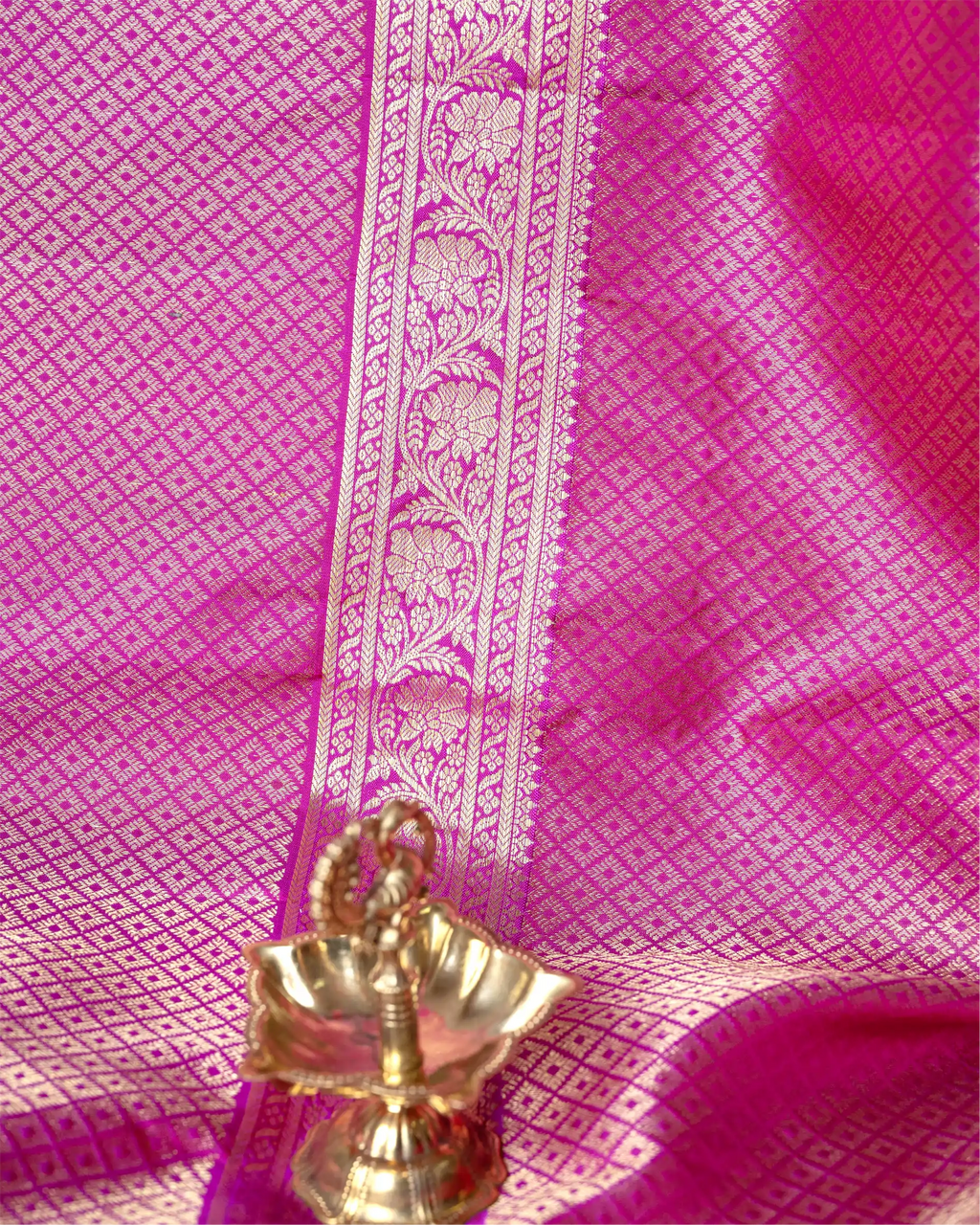 Close-up of a bright pink Kanjivaram silk saree fabric featuring a detailed gold and silver zari floral woven border with a small brass decorative lamp in the foreground.