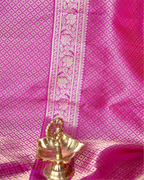 Close-up of a bright pink Kanjivaram silk saree fabric featuring a detailed gold and silver zari floral woven border with a small brass decorative lamp in the foreground.
