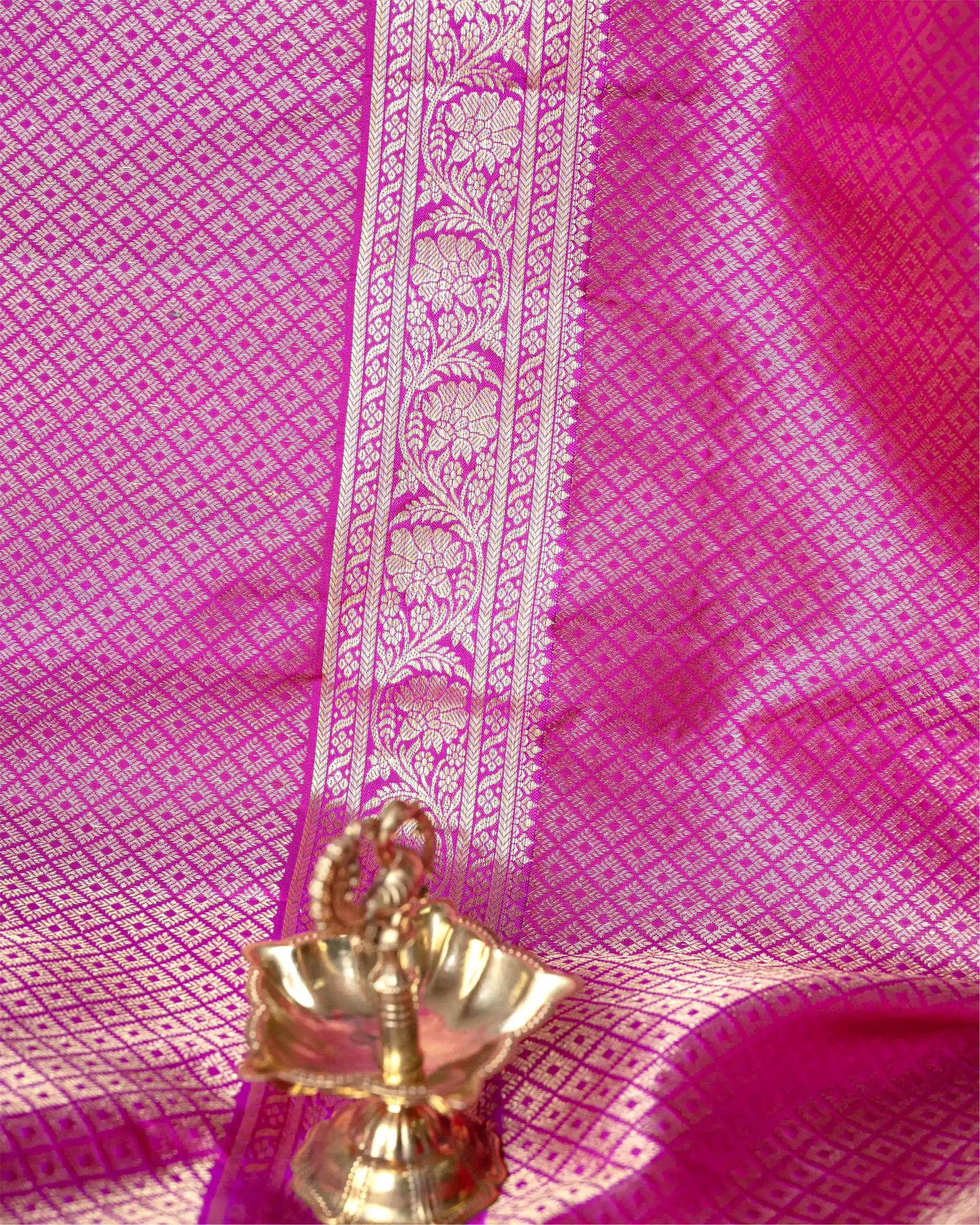 Close-up of a bright pink Kanjivaram silk saree fabric featuring a detailed gold and silver zari floral woven border with a small brass decorative lamp in the foreground.