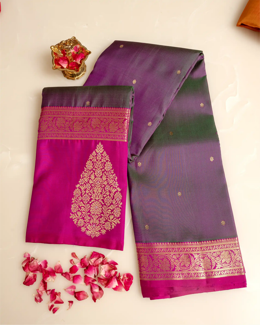 A folded dark green and magenta Kanjivaram silk saree with an ornate gold zari brocade pallu and woven borders, displayed on a beige surface with scattered pink rose petals and a small decorative pot