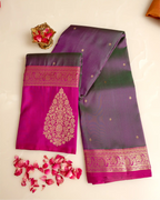 A folded dark green and magenta Kanjivaram silk saree with an ornate gold zari brocade pallu and woven borders, displayed on a beige surface with scattered pink rose petals and a small decorative pot
