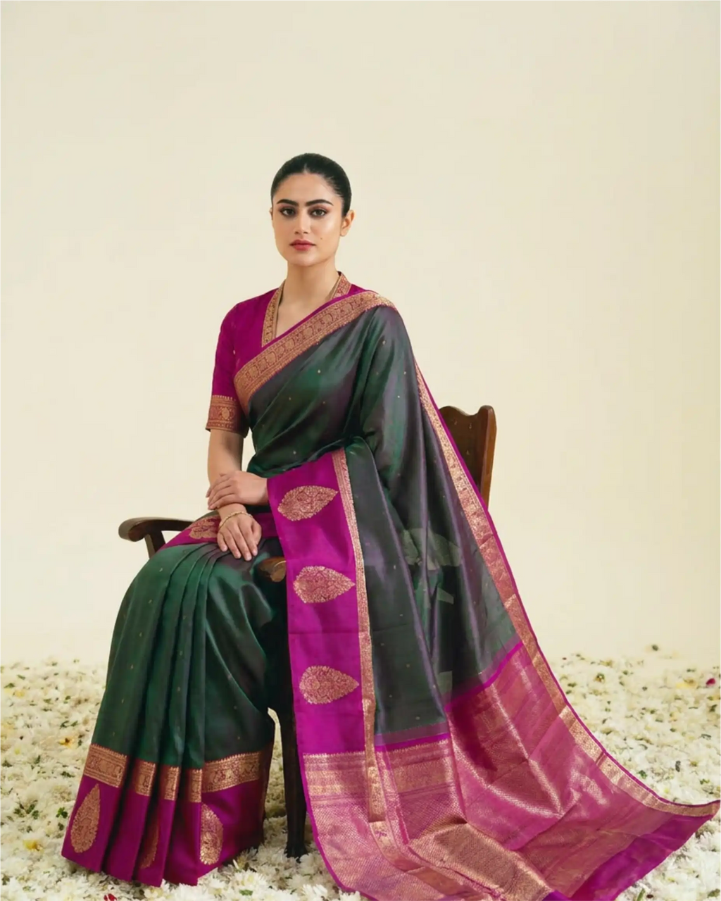 A woman seated on a wooden chair wearing a dark green and magenta silk saree with gold zari borders and motifs, set against a plain cream backdrop with scattered white flower petals on the floor.