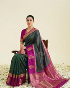 A woman seated on a wooden chair wearing a dark green and magenta silk saree with gold zari borders and motifs, set against a plain cream backdrop with scattered white flower petals on the floor.