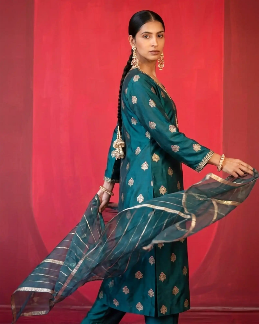 A model standing against a red backdrop wearing a premium dark teal pure Banarasi silk suit with gold weaving and a matching sheer Kora dupatta.