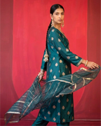 A model standing against a red backdrop wearing a premium dark teal pure Banarasi silk suit with gold weaving and a matching sheer Kora dupatta.