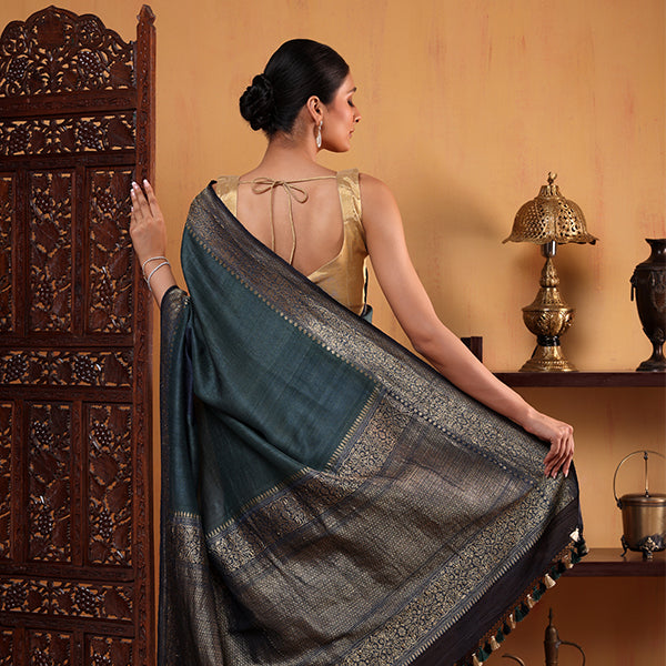 Back view of a woman wearing a luxurious deep teal Banarasi silk saree, featuring exquisite gold zari weaving on the pallu and traditional patterns, paired with a matching gold sleeveless blouse.