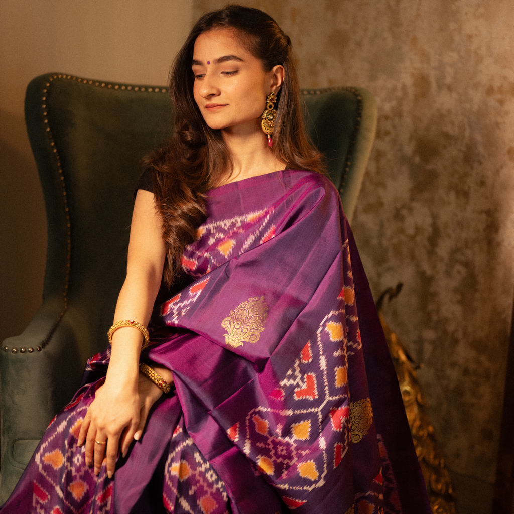 Woman in a purple saree with intricate patterns sitting on a green chair.