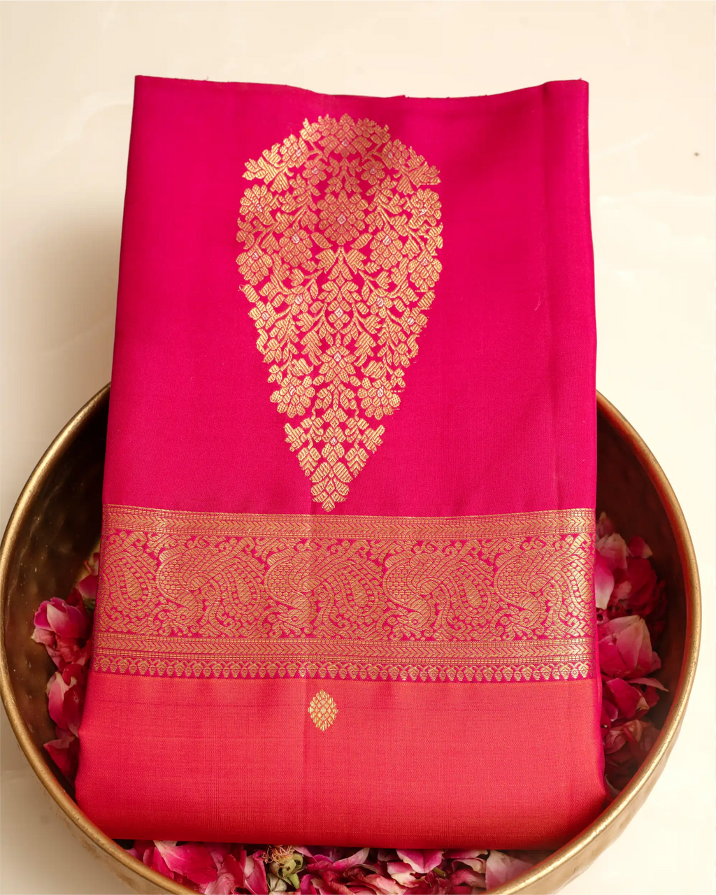 A folded bright pink Kanjivaram silk saree with an intricate gold zari woven border, displayed in a brass bowl filled with fresh rose petals