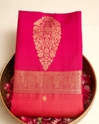 A folded bright pink Kanjivaram silk saree with an intricate gold zari woven border, displayed in a brass bowl filled with fresh rose petals