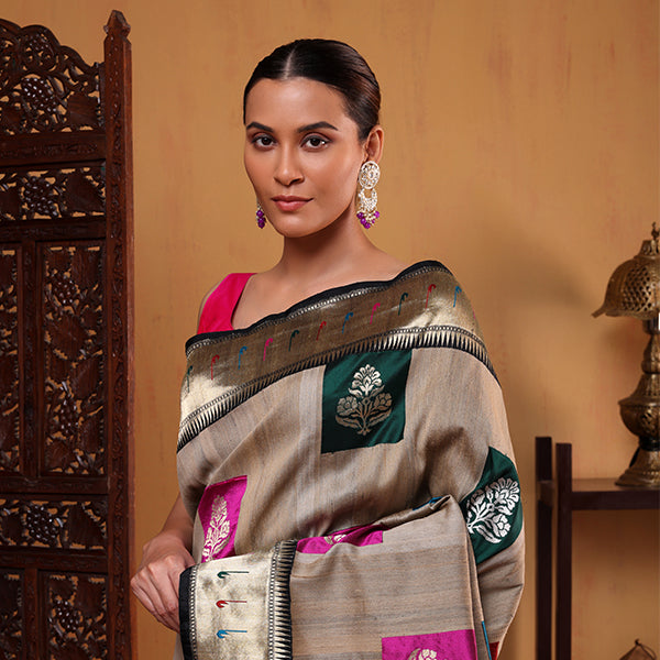 A woman posing against a wooden screen, elegantly draped in a golden beige pure Tussar silk saree with multicolored zari bootas and a heavy border.
