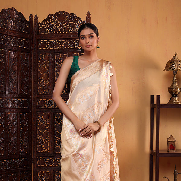 A woman posing in a beautifully decorated room, wearing an elegant beige tissue silk saree with intricate golden zari work and a contrasting sleeveless green blouse.
