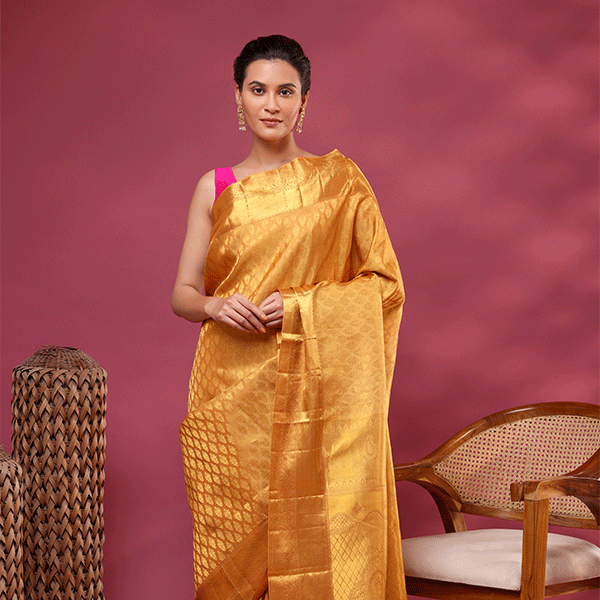 A woman posing against a dusty pink background, elegantly draped in a golden mustard yellow Kanjivaram silk saree with intricate resham zari work, paired with a vibrant pink sleeveless blouse.