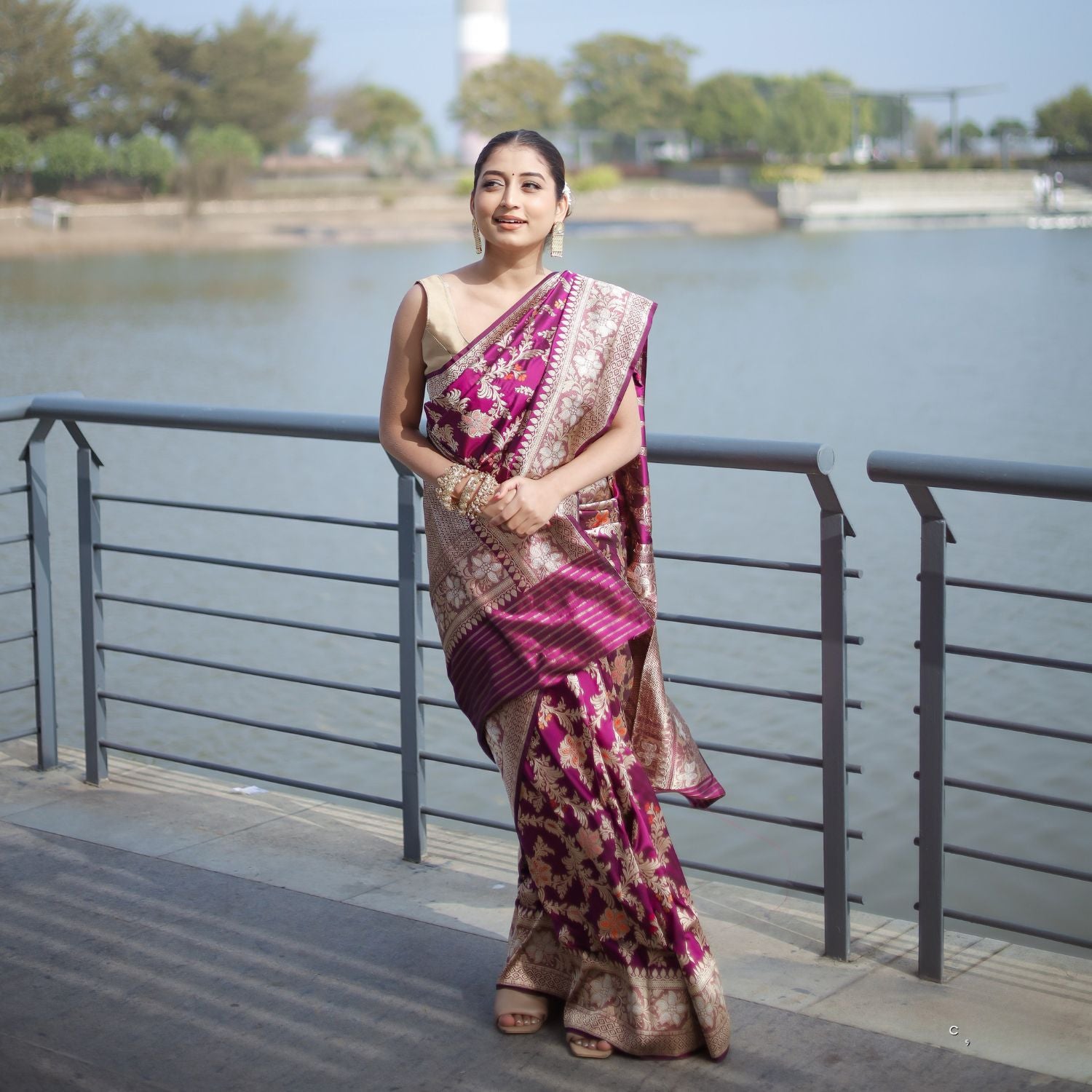 A woman standing on a bridge wearing a traditional, handwoven katan silk saree in purple and silver colors with a floral pattern.