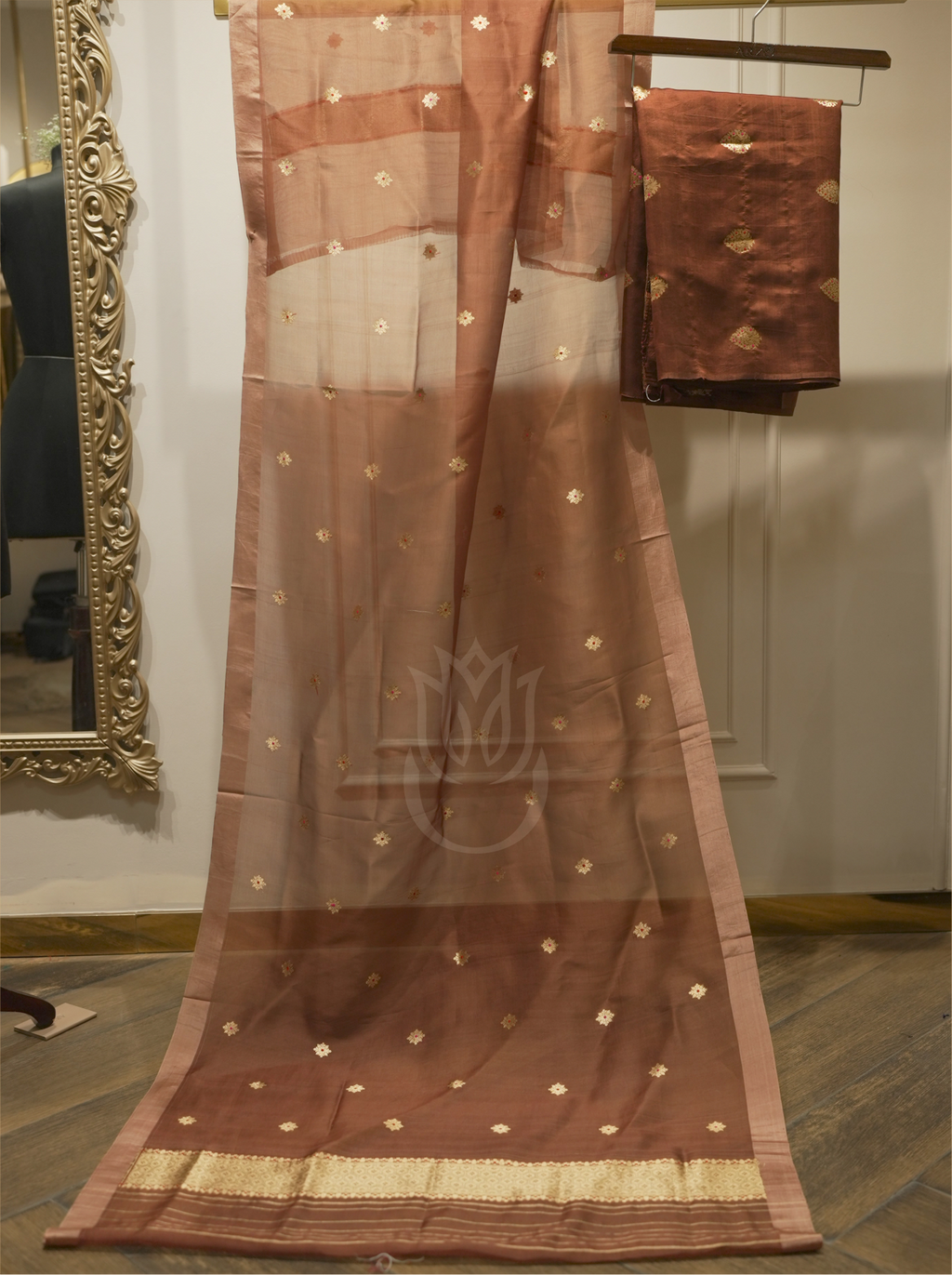 Sheer chocolate brown handwoven Banarasi Kora silk dupatta with starburst zari motifs displayed vertically alongside folded silk suit fabric on a wooden hanger, set near a gold ornate mirror