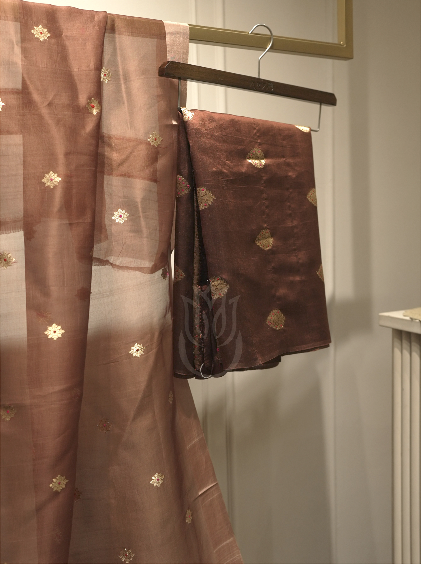 Chocolate brown handwoven Banarasi Kora silk dupatta with starburst gold zari motifs displayed vertically alongside folded suit fabric on a wooden hanger, set near a gold ornate mirror
