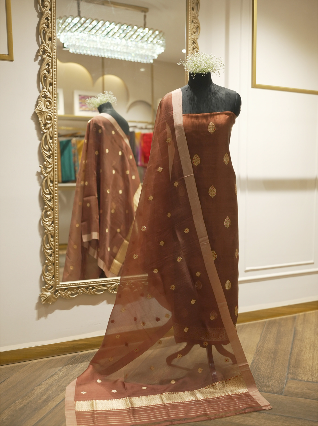 Full-length display of a chocolate brown handwoven Banarasi Kora silk suit on a mannequin torso, styled with a sheer gold zari starburst dupatta reflected in an ornate gold mirror