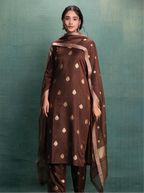 Woman modeling a chocolate brown handwoven Banarasi silk kurta adorned with gold zari kalga bootis, styled with a draped sheer Kora silk dupatta featuring zari borders against a textured green background