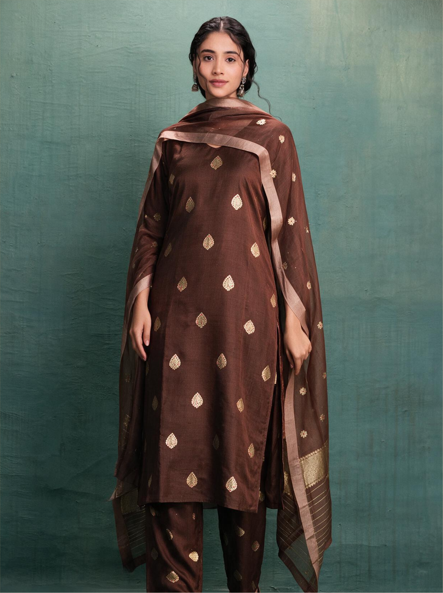 Woman modeling a chocolate brown handwoven Banarasi silk kurta adorned with gold zari kalga bootis, styled with a draped sheer Kora silk dupatta featuring zari borders against a textured green background