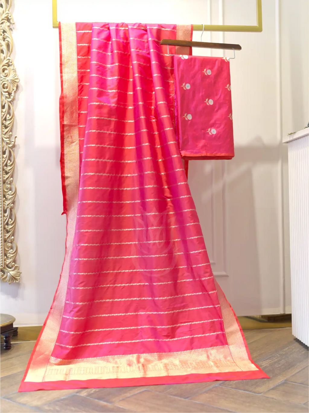 Handwoven magenta pink Banarasi tissue dupatta with horizontal gold zari stripes and a woven border draped on a gold stand next to a matching silk suit piece with silver floral bootis.