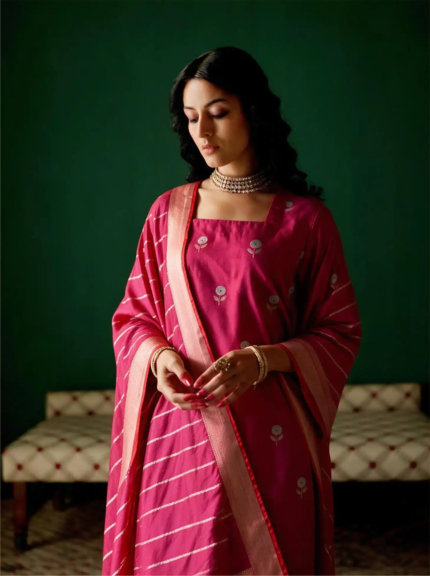 Woman posing in a handwoven magenta pink Banarasi silk suit set featuring silver floral bootis and a tissue zari striped dupatta against a dark green backdrop.