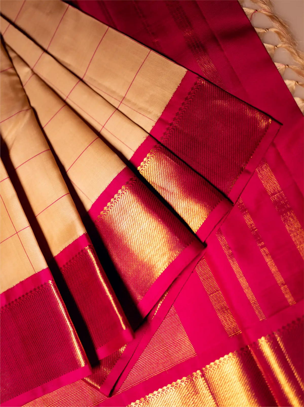 Close-up view of a folded handwoven silk saree featuring a beige checked body with shimmering gold zari lines and a contrasting vibrant magenta-pink border with metallic gold zari weave.