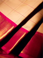 Diagonal close-up shot of a handwoven silk saree in beige and gold with subtle checked grid lines, featuring a rich crimson red border with fine golden zari weaving along the folded edges.