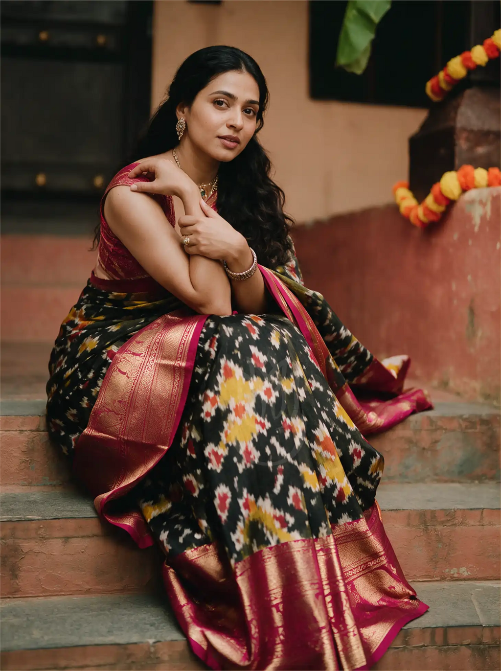 A woman sitting on wooden steps modeling a handwoven black Kanji Patola silk saree featuring vibrant geometric Patola motifs in red, yellow, and green, complete with a rich crimson red zari border.