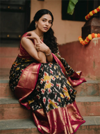 A woman sitting on wooden steps modeling a handwoven black Kanji Patola silk saree featuring vibrant geometric Patola motifs in red, yellow, and green, complete with a rich crimson red zari border.